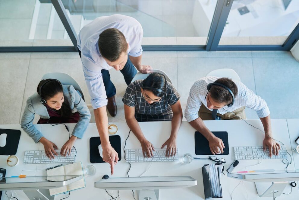 A supervisor is pointing over the shoulder of a worker at the monitor screen, while two other employees look at where the supervisor is pointing.